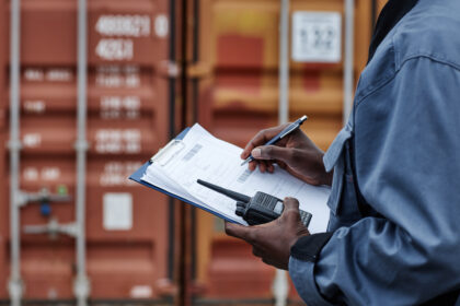 Close up of male worker wearing writing on clipboard in shipping docks with containers, copy space